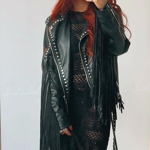 Könnte beinhalten: Schwarze Lederjacke mit silbernen Nieten und langen Fransen. Die Jacke hat einen Reißverschluss und einen Stehkragen.