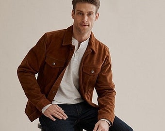 May include: A man wearing a brown suede jacket, a white long-sleeved shirt, dark blue jeans, and brown suede ankle boots. He is sitting on a black stool.