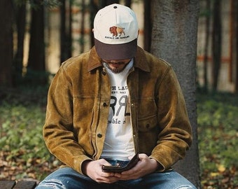 May include: A person wearing a brown suede jacket, blue jeans, and brown boots sits on a stone wall. They are wearing a white baseball cap with a brown buffalo graphic and the text "Wrangler" on the front. They are looking down at their phone.