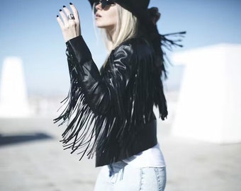 May include: A woman wearing a black leather jacket with fringe detail, a black hat, and blue jeans.