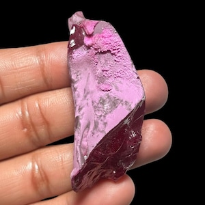 May include: A vibrant, irregularly shaped gemstone with a striking color contrast. The top half is a light, textured pink, while the bottom half is a deep, translucent burgundy. The gemstone is held against a dark background, highlighting its unique form.