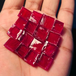 May include: A square mosaic of small, ruby-red glass tiles. The tiles are arranged in a grid pattern, with each tile reflecting light. The mosaic is held in a hand, showcasing the vibrant color and texture of the glass.