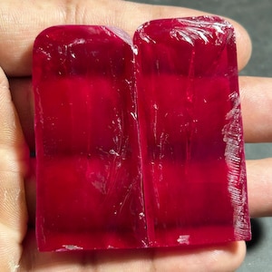 Puede incluir: Dos piedras preciosas rectangulares de rubí rojo intenso con la parte superior redondeada. Las gemas tienen una calidad translúcida, con fracturas internas visibles y una superficie ligeramente rugosa e irregular. Las piedras se sostienen en una mano, mostrando su rico color y textura natural.