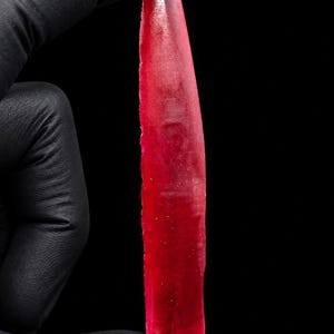Puede incluir: Un espécimen de cristal rojo largo, delgado y translúcido con una punta afilada y una base plana. La piedra preciosa exhibe un tono carmesí profundo con texturas superficiales naturales y variaciones internas visibles en su estructura.