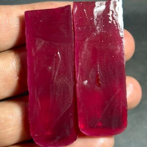 May include: Two raw, rectangular ruby gemstones with a deep red hue. The stones have a translucent quality, with visible internal structures and a slightly uneven surface. The edges are rough, indicating they are uncut and natural.