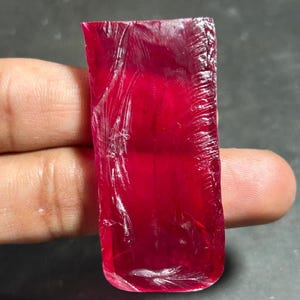May include: A raw, rectangular ruby gemstone with a deep red hue. The surface of the stone has visible fractures and imperfections. The gemstone is held up against a dark background.