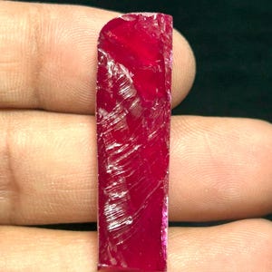 May include: A raw, rectangular ruby gemstone with a deep red hue. The surface of the stone has visible fractures and a slightly polished appearance. The ruby is held in a hand, showcasing its natural, unrefined form.