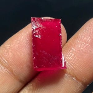 Pode incluir: Uma gema retangular, translúcida, vermelho escuro, com uma superfície ligeiramente áspera e texturizada. A gema é segurada entre os dedos contra um fundo escuro, mostrando sua cor vibrante e estrutura cristalina. A gema parece ser uma rubi.