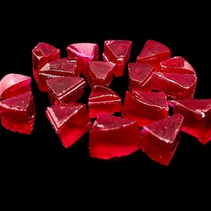 Op de afbeelding: Een verzameling kleine, doorschijnende, dieprode edelsteenfragmenten met onregelmatige, hoekige vormen en gefacetteerde oppervlakken. Deze stukken vertonen een levendige karmozijnrode tint en een glasachtige textuur.