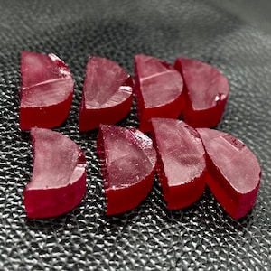 Puede incluir: Un grupo de ocho piezas de caramelo rojo, semicirculares y translúcidas, dispuestas en forma de semicírculo sobre una superficie negra texturizada.