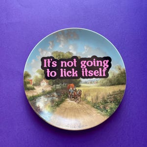 May include: A round decorative plate featuring a pastoral landscape painting of a horse-drawn carriage on a dirt path near a stream. The text "It's not going to lick itself" is printed in bold pink letters across the center of the scene.
