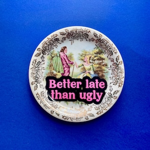 May include: A round ceramic trinket dish featuring a gold floral border and a central illustration of two figures in period clothing. A black sticker with pink text reading Better late than ugly is placed over the center of the dish.