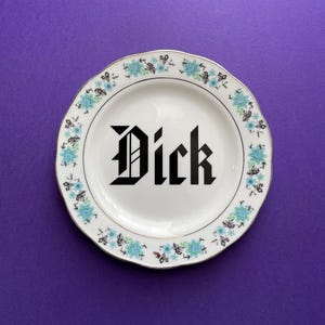 May include: A white ceramic plate with a blue floral border. The word "Dick" is written in black block letters in the center of the plate.
