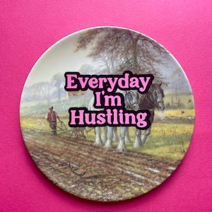 May include: A round ceramic decorative plate featuring a pastoral scene of a farmer plowing a field with two draft horses. The text "Everyday I'm Hustling" is printed in bold pink letters across the centre of the image over the rural landscape.