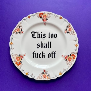May include: A white ceramic plate with a scalloped edge featuring a floral pattern in orange, yellow, and grey. The center of the plate displays the black text "This too shall fuck off" written in a gothic-style font with decorative flourishes.