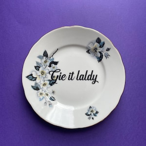 May include: White ceramic plate with a gold rim and floral design. The plate has the text "Gie it laldy" written in black.