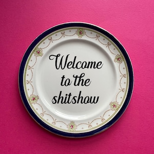May include: A white ceramic plate with a blue and gold rim. The plate has a floral design and the text "Welcome to the shitshow" written in black.