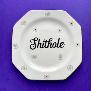 May include: White octagonal ceramic plate with a gold rim and black starburst designs around the edge. The plate has the word "ShitHole" written in black cursive lettering in the center.