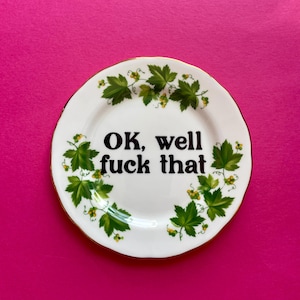 May include: A round white ceramic plate with a gold-trimmed edge, featuring a decorative border of green grape leaves and small yellow clusters. The center of the plate displays the black text "OK, well fuck that" in a bold, serif typeface.
