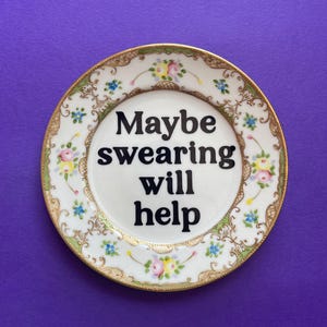 May include: A round white ceramic plate featuring the black text Maybe swearing will help in the center. The rim is decorated with a gold-trimmed edge and a pattern of small pink, yellow, and blue flowers with green leaves on a white background.