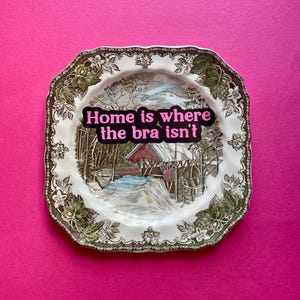 May include: A square ceramic plate with scalloped edges featuring a brown and white transferware landscape of a covered bridge. The center of the plate displays a black sticker with the pink text "Home is where the bra isn't" in a bold font.
