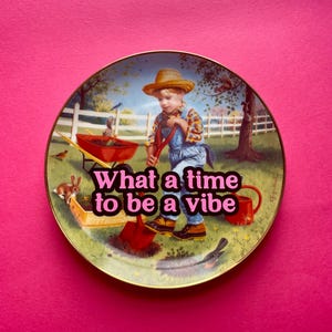 May include: A round decorative plate with a gold-toned rim featuring a printed illustration of a child in a straw hat and denim overalls gardening with a red shovel. The text "What a time to be a vibe" is printed in pink across the center.