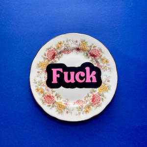 May include: A round white ceramic plate featuring a decorative border of pink and yellow floral patterns with green leaves. Centered on the plate is a black rectangular sticker with the word "Fuck" printed in bold, bright pink capital letters.