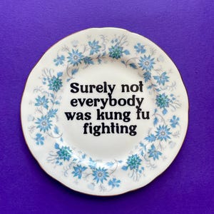 May include: A white ceramic plate with a scalloped gold-trimmed edge, featuring a blue and green floral pattern around the rim. The center of the plate displays the black text: Surely not everybody was kung fu fighting, written in a bold serif font.