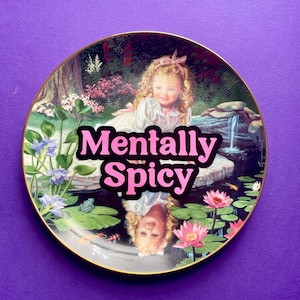 May include: A round decorative plate with a gold rim featuring a printed illustration of a blonde child in a garden setting by a pond. The text Mentally Spicy is printed in bold pink letters across the center of the image over the child.