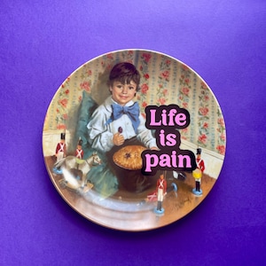 May include: A round decorative plate featuring a vintage-style illustration of a child in a white shirt and blue bow tie sitting with a pie and toy soldiers. The text "Life is pain" is printed in bold pink letters across the center of the plate.