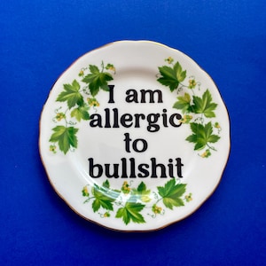 May include: A round white ceramic plate with a gold-trimmed scalloped edge. The center features the black text "I am allergic to bullshit" surrounded by a decorative border of green grape leaves and small yellow clusters of grapes.