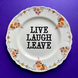 May include: White ceramic plate with floral accents in orange, yellow, and gray. The plate features the words "LIVE LAUGH LEAVE" in bold black letters. The plate has a scalloped edge and is set against a purple background.