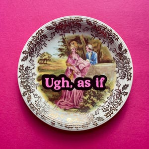 May include: A round ceramic decorative plate featuring a central illustration of a person in a pink gown and another in a blue coat within a landscape. The rim has a gold floral pattern. The text Ugh, as if is printed in pink over the center.
