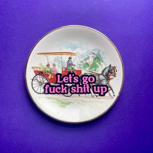 May include: A round white ceramic decorative plate with a gold-toned rim. The center features a vintage-style illustration of a horse-drawn carriage with passengers and the bold pink text "Let's go fuck shit up" layered over the scene.