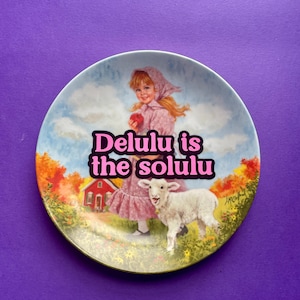 May include: A round decorative ceramic plate featuring a printed illustration of a young person in a pink dress and bonnet holding a red apple, standing next to a white lamb. The text "Delulu is the solulu" is printed in pink with a black outline.