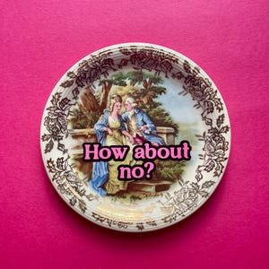 May include: A round ceramic decorative plate featuring a central illustration of two figures in period clothing sitting on a stone wall. The plate has a brown floral border and the text How about no? in bold pink letters across the center.