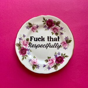 May include: A white ceramic saucer with a scalloped gold-trimmed edge, decorated with a floral pattern of pink and dark red roses and small purple flowers. The center features the black text "Fuck that Respectfully" in a bold, serif font.