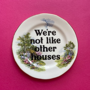 May include: A round white ceramic plate with a gold-trimmed edge. The center features the black text "We're not like other houses" surrounded by a colorful illustration of a cottage, green trees, and blooming flowers on both sides.