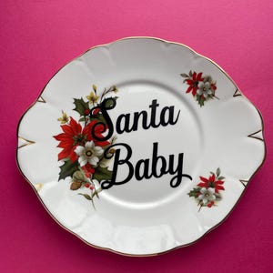 May include: White ceramic plate with a gold rim and a festive design. The plate features red and white poinsettia flowers, green holly leaves, and the text "Santa Baby" in black lettering.