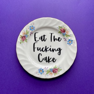 May include: White ceramic plate with gold trim and floral accents. The plate features the text "Eat The Fucking Cake" in black script. The plate is set against a purple background.
