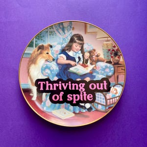 May include: A round decorative plate with a gold rim featuring an illustration of a child reading to a collie dog and a teddy bear while sitting in a blue floral armchair. The text "Thriving out of spite" is printed in pink over the center.