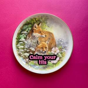 May include: A round white ceramic decorative plate with a gold-trimmed edge featuring a detailed illustration of two foxes nestled among green foliage and white flowers. The text "Calm your tits" is printed in bold pink letters across the center.