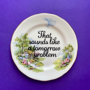 May include: A white ceramic plate with a gold-trimmed edge featuring a colorful landscape illustration of a cottage, trees, and flowers. The center of the plate displays the black text, That sounds like a tomorrow problem, in a cursive font.