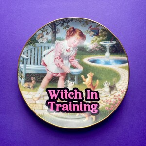 May include: A round decorative ceramic plate with a gold rim featuring a garden scene with a child in a pink dress and white apron feeding kittens. The text "Witch In Training" is printed in bold pink letters across the center of the illustration.