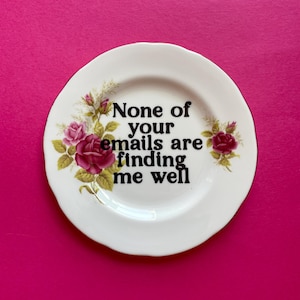 May include: A white ceramic plate with a scalloped edge featuring a floral design of pink roses and green leaves on the left and right sides. The center of the plate displays the black text: None of your emails are finding me well.