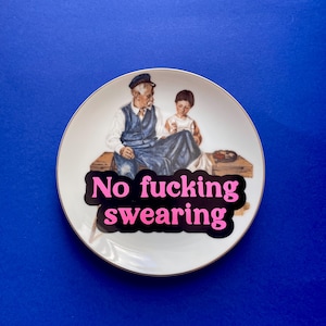 May include: A white ceramic decorative plate with a gold-trimmed edge featuring an illustration of an older person and a child sitting on a wooden bench. Overlaid on the image is a black speech bubble shape with the pink text No fucking swearing.