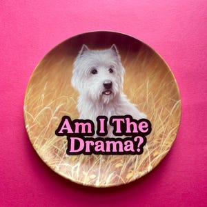 May include: A round decorative plate featuring a portrait of a white West Highland White Terrier dog sitting in a field of tall golden grass. The text "Am I The Drama?" is printed in bold pink letters with a black outline across the center.