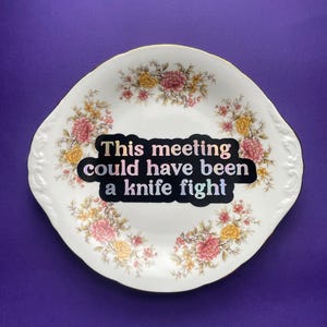 May include: A white ceramic plate with a scalloped edge and gold trim, decorated with a floral pattern of pink and yellow flowers. A black sticker in the center features the text: This meeting could have been a knife fight.