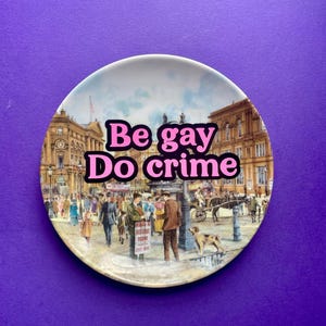 May include: A round ceramic decorative plate featuring a vintage-style illustration of a busy city square with people, buildings, and a horse-drawn carriage. The text "Be gay Do crime" is printed in bold pink letters across the center of the image.