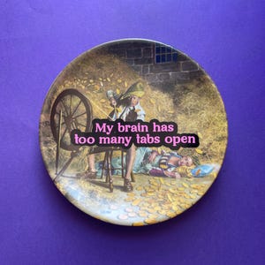 May include: A round decorative plate featuring a classic illustration of a person spinning straw into gold coins. The text 'My brain has too many tabs open' is printed in pink letters across the center of the image, overlaying the scene.